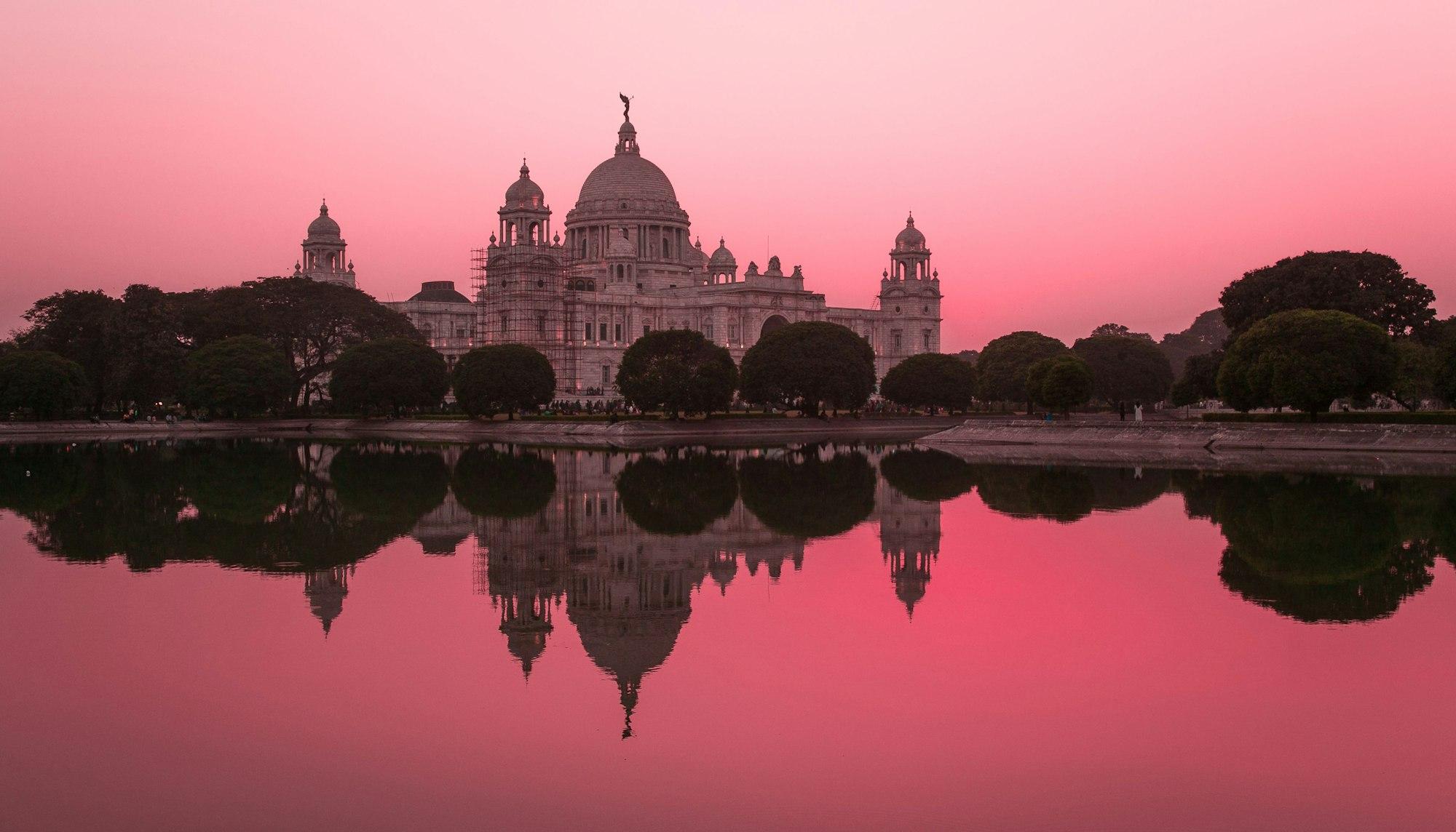 The domed Victoria Memorial in Kolkata reflected in still water under a pink sky